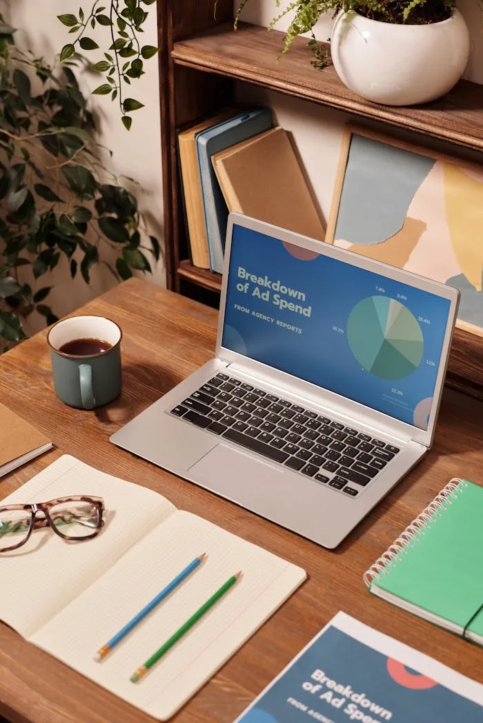 Laptop displaying ad spend data on a wooden desk with office supplies.